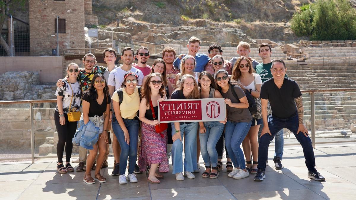 a large group of Trinity students and faculty pose with a banner in front of an historic site in Madrid, Spain