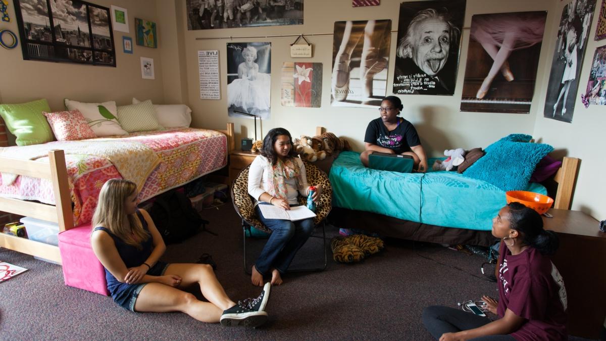 Four students, one sitting on a bed and the other three sitting on the floor in their dorm room having a conversations